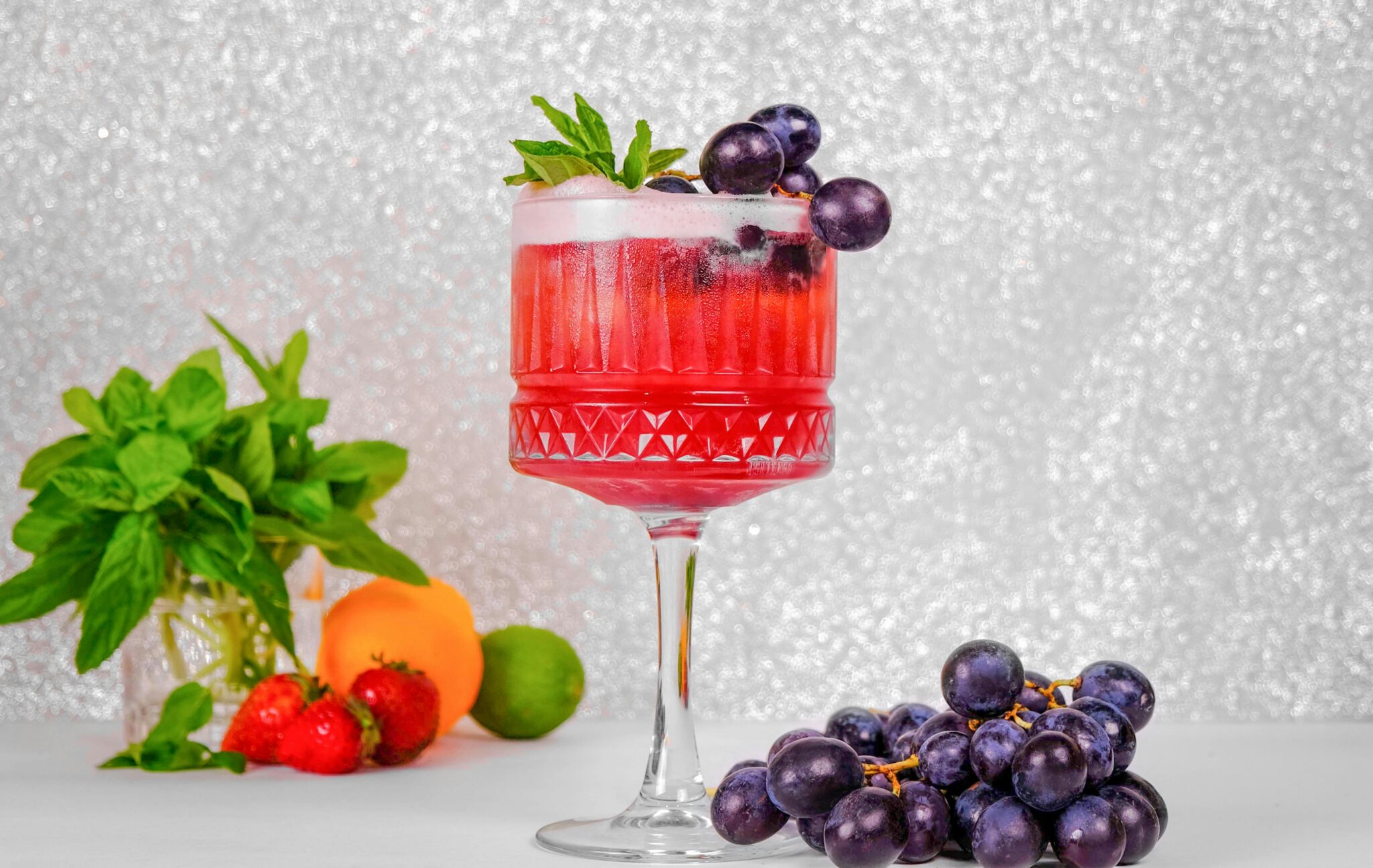 Delicious red cocktail garnished with grapes and mint, surrounded by fresh fruits.